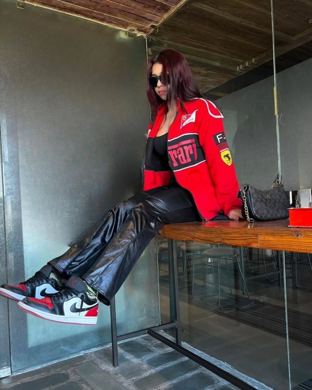 Female model sitting in a red Ferrari bomber jacket, paired with black leather pants and Jordan sneakers, styled for urban F1 apparel fashion.