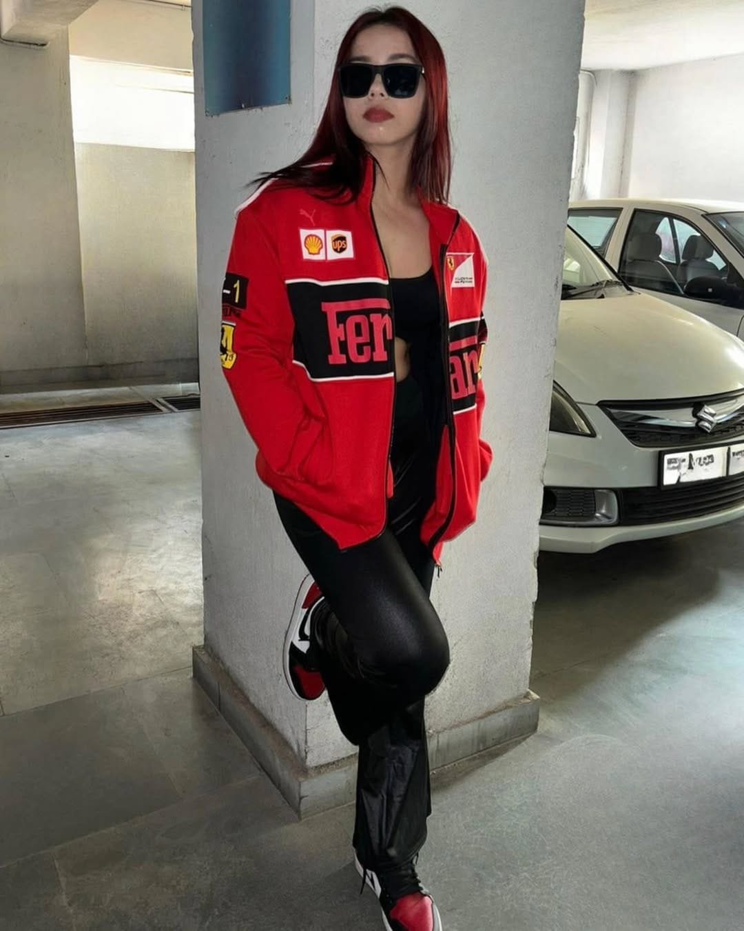 Woman posing in parking area wearing red Ferrari racing jacket, black leather pants, and sneakers, representing premium F1 apparel by Kezual.